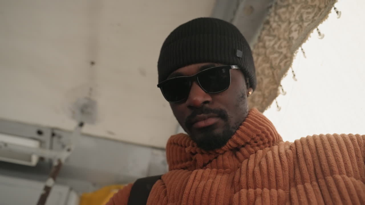 Uomo di colore con berretto che controlla il telefono, maglione a coste arancione e occhiali da sole, primo piano dal basso all'interno di un autobus urbano, umore riflessivo, tragitto mattutino, stile street candid, interazione con lo smartphone, toni smorzati