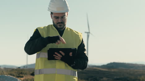 promoting sustainable energy sources, a professional engineer in a white helmet and reflective vest uses a tablet to audit wind turbines on a sunny day in a field of renewable energy generators