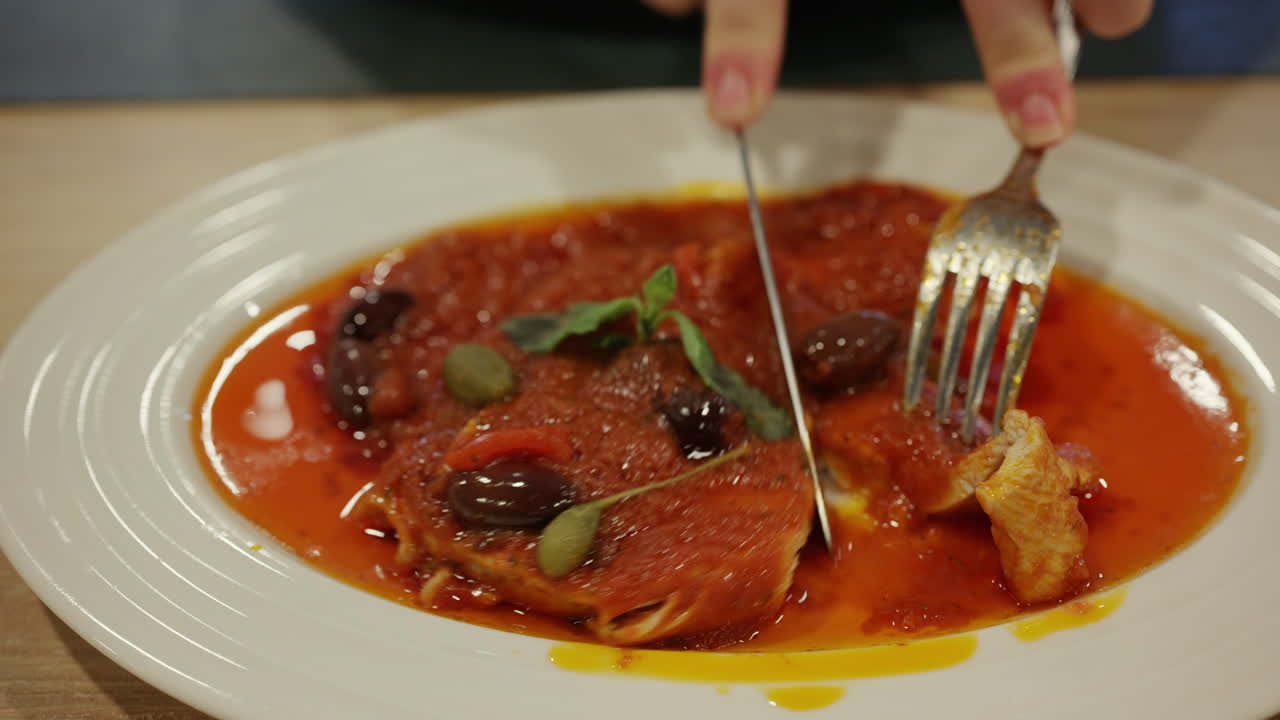 Primo piano di pollo in un ricco sugo di pomodoro con olive e capperi, tagliato con forchetta e coltello