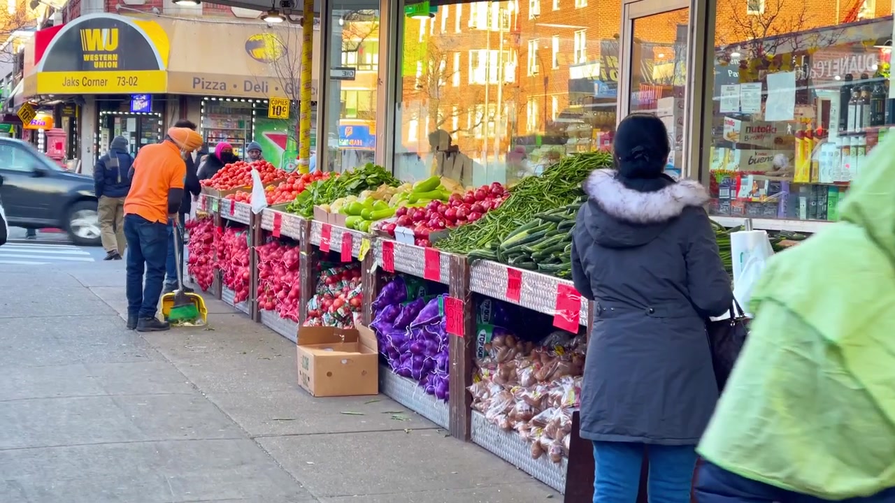 Nueva York, EE. UU., 9 de marzo de 2022: Puesto de frutas y verduras en la acera con un comprador en Jackson Heights. Vista amplia desde la acera de una exhibición de verduras al aire libre con un comprador, reflejos de escaparates y luz de la tarde.