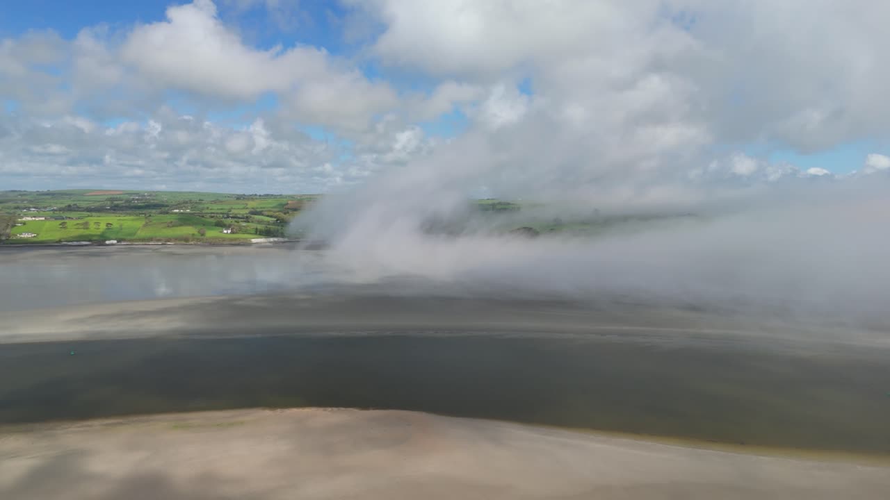 Mist is lifting and creating clouds over water and green hills, drone footage
