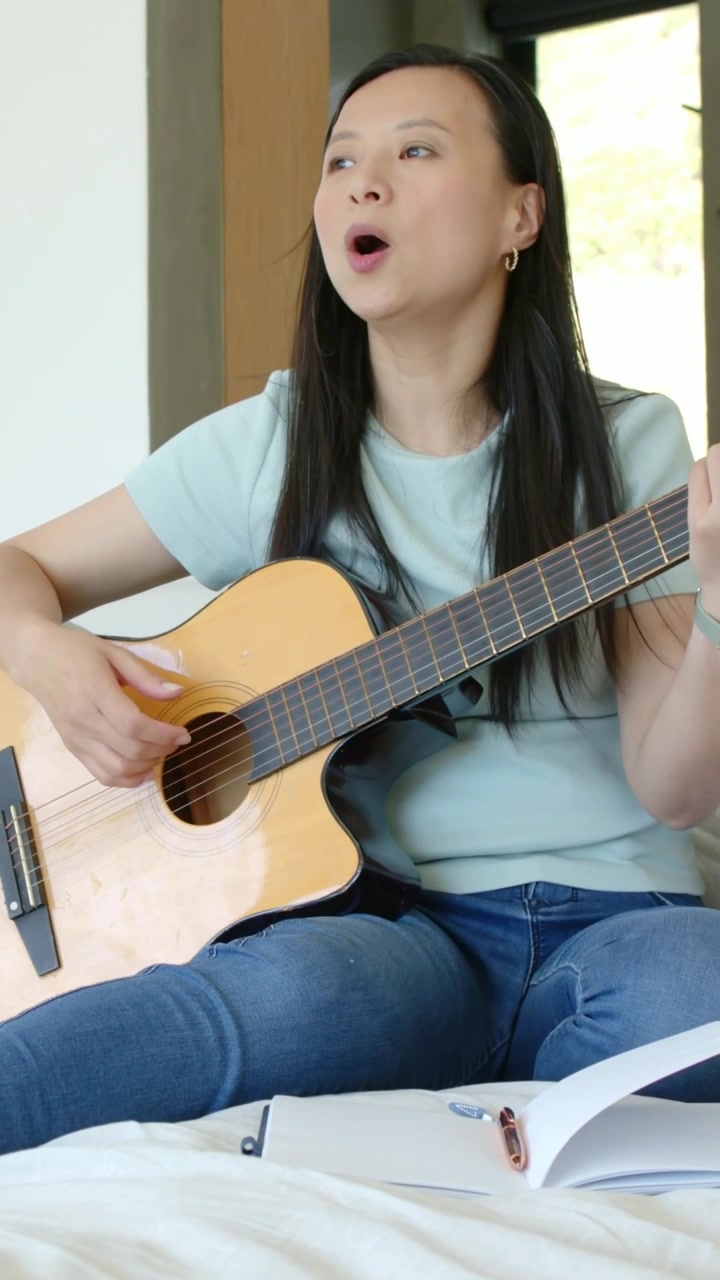 Video verticale: Donna in maglietta blu che inizia a strimpellare la chitarra sul letto vicino alla finestra, controllando gli appunti.