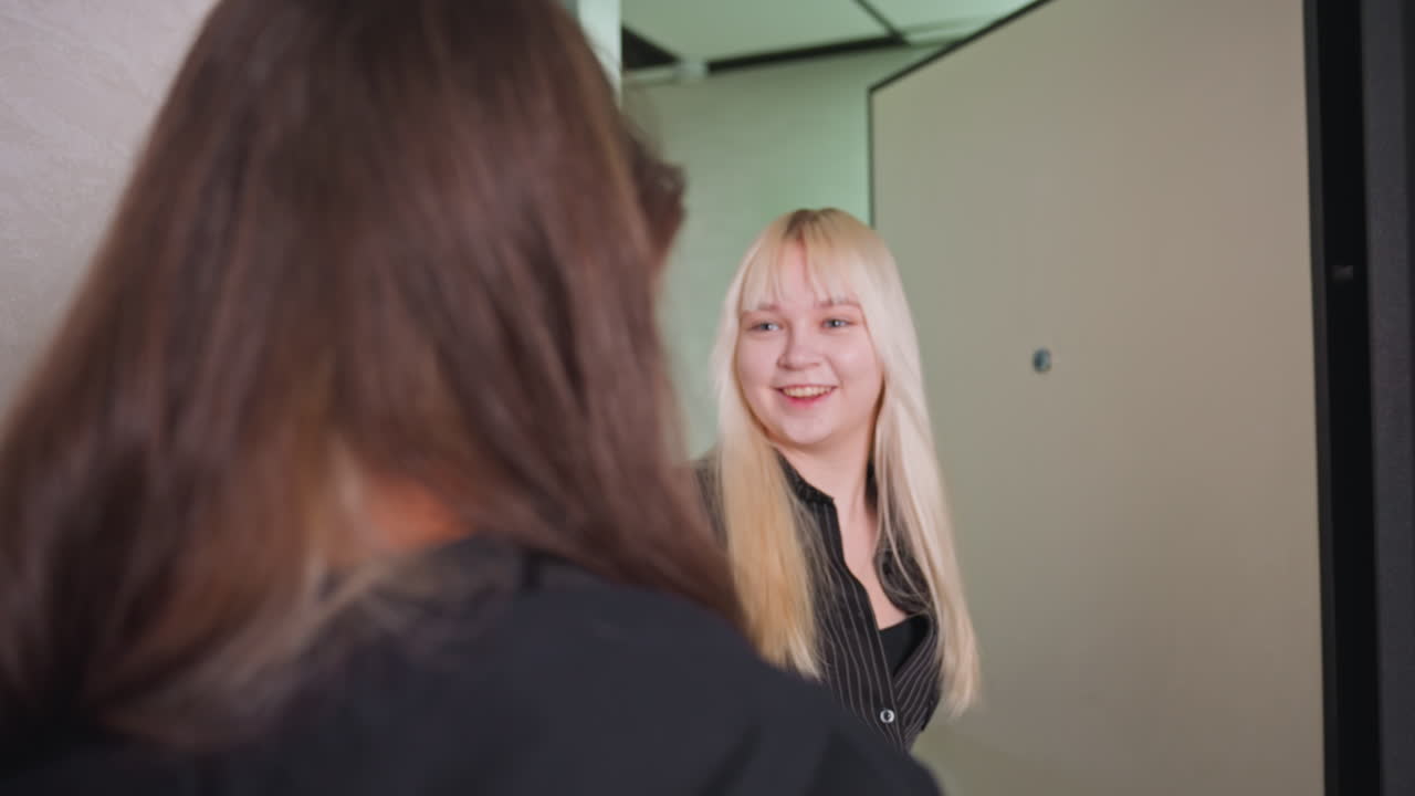 Rear view of woman in black hoodie opening door for friend and sharing warm hug in modern hallway, then friend drops her bag on floor as they embrace