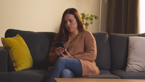 woman sitting on sofa at home using mobile phone to check social media message and read news article