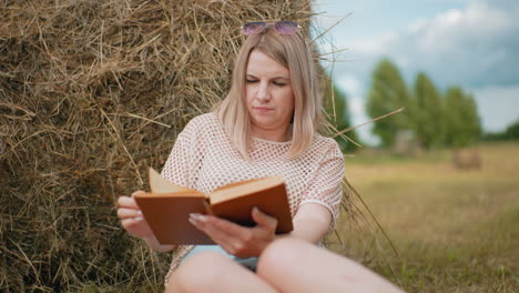 woman sitting with legs joined, deeply engrossed in reading while leaning against hay bale, thoughtful expression as she flips pages, with soft blur in the background of trees and open countryside