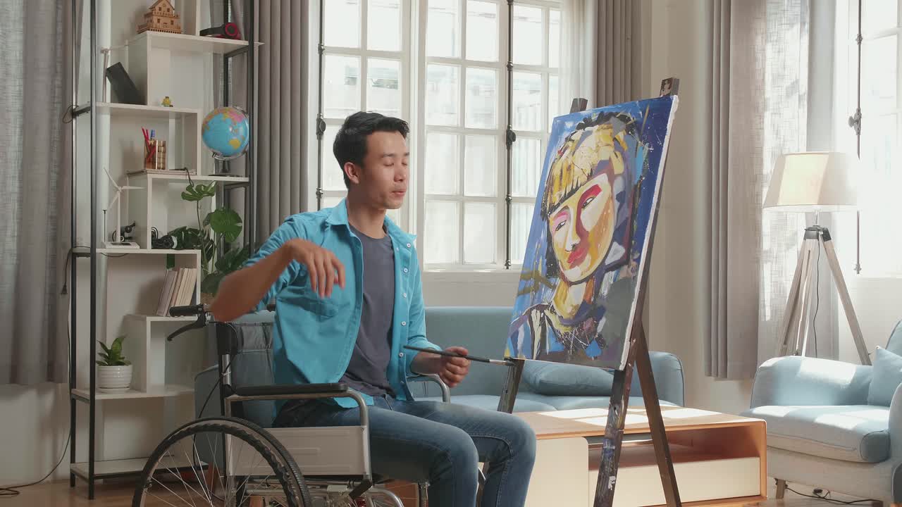 vista trasera de un artista asiático hombre en silla de ruedas sosteniendo un pincel gira para limpiar el sudor antes de cruzar los brazos y sonriendo mientras pinta una chica en el lienzo