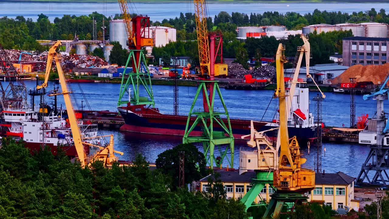 Industrial port scene with multiple cargo cranes, docked freight ships, stacked materials, and surrounding infrastructure, captured from above in vibrant daylight