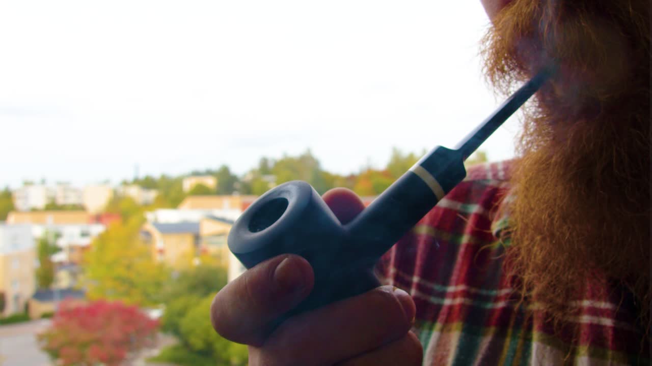 primer plano de un hombre moderno de barba roja sosteniendo una pipa antigua de color negro oscuro en su mano y fumando al aire libre en su balcón con una camisa roja soplando tabaco de calidad casas de día nublado