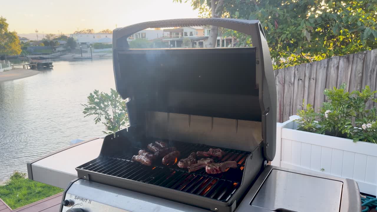 Sausages and lamb cutlets sizzle with flames and smoke on a gas grill at sunset