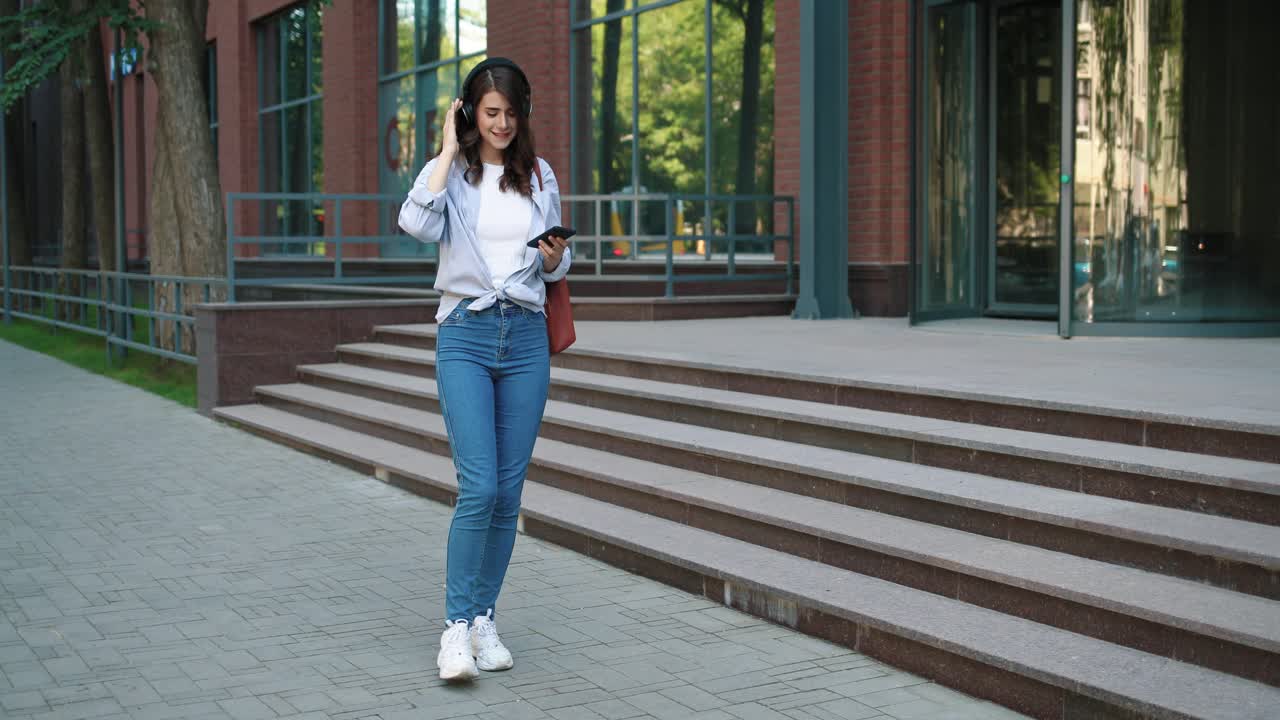 Full length view of caucasian student woman wearing headphones and listening to the music on smartphone while walking down the street