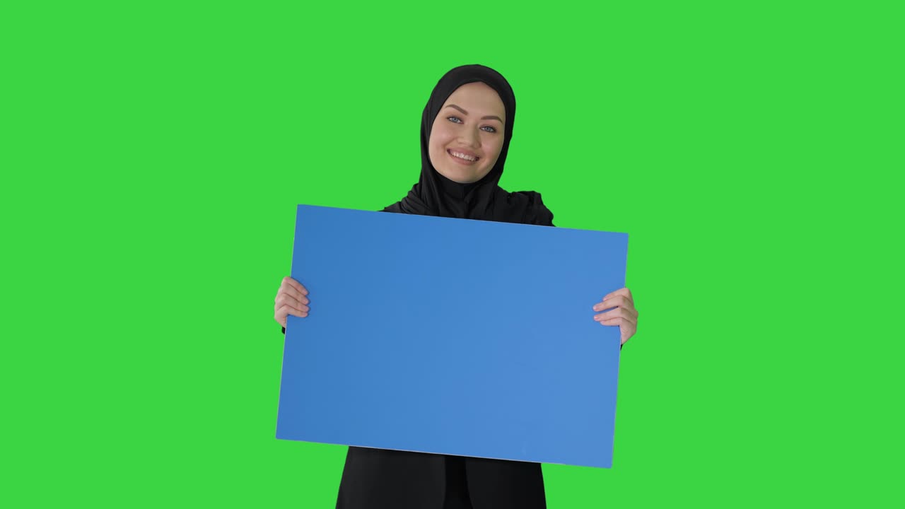 Donna araba sorridente con hijab che tiene in mano un poster blu vuoto e lo guarda su schermo verde, Chroma Key