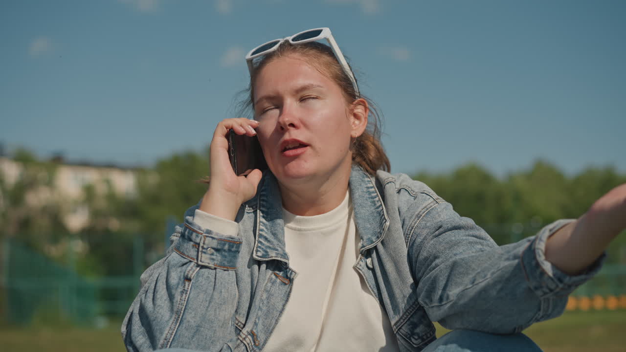 Jonge vrouw geanimeerd aan de telefoon buiten, spijkerjack en zonnebril, expressieve handgebaren, hek van sportveld op de achtergrond, helderblauwe lucht, openhartige vertellerenergie en dynamische vertelling