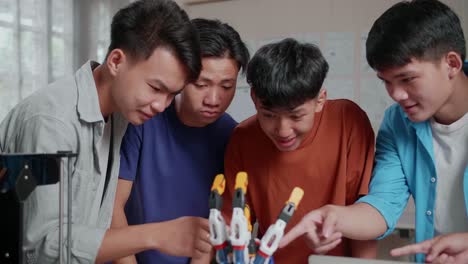 grupo de jóvenes chicos asiáticos con una computadora portátil e impresión 3d sonriendo y mostrando los pulgares hacia arriba gesto a la cámara mientras discuten sobre una mano cyborg en casa
