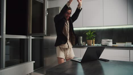 A-brunette-man-with-stubble-in-a-black-jacket-and-beige-shorts-rejoices-at-his-success-and-has-fun-while-working-remotely-in-the-kitchen-in-a-modern-apartment-in-the-evening