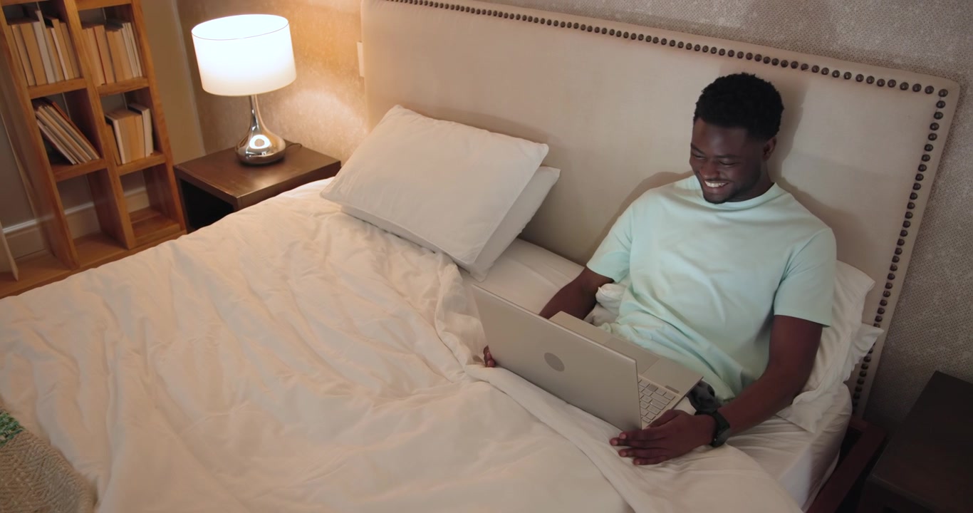 African American man reacting to on-screen content, laughing and typing on laptop on bed