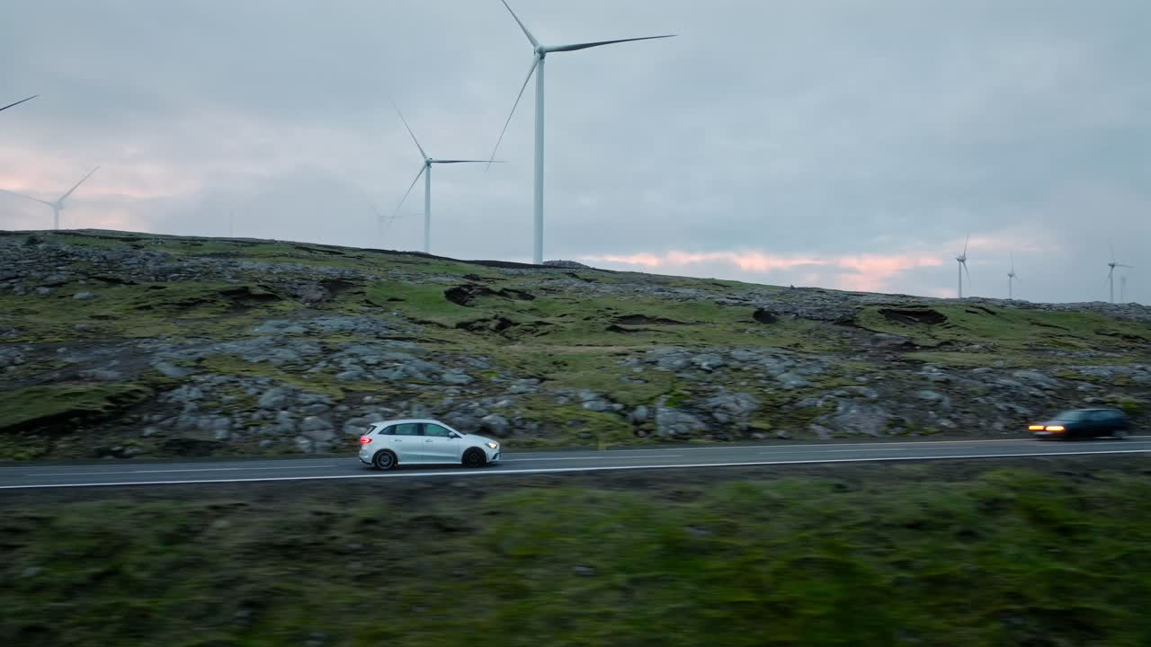 Two cars drive on a rural road with wind turbines spinning on a cloudy day