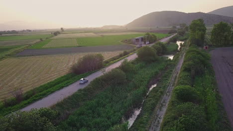 Aerial-shot-of-car-driving-down-country-road-surrounded-by-fields,-4K