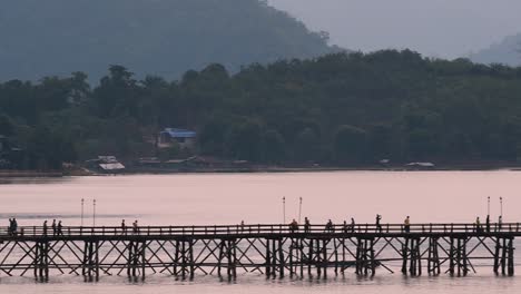 Mon-Bridge-and-a-longboat-followed-speeding-behind-the-structure-while-people-are-walking-on-the-bridge,-silhouetting-as-it-was-getting-dark,-in-slow-motion