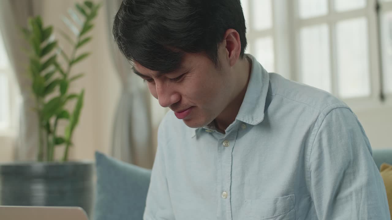 Asian Male Working On Laptop And Smiling At Living Room While Sitting On Sofa, Video In 4K