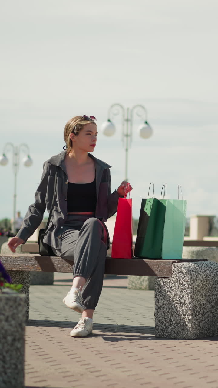 한 여인이 야외 벤치에 앉아 쇼핑백을 내려놓고 빨간색 쇼핑을 조정하고 벤치 위에 손을 고 편안하게 앉아 공원 벤치와 꽃으로 둘러싸인 오른쪽을 바라본다.