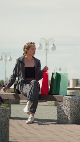 la señora se sienta en un banco al aire libre, deja caer sus bolsas de compras, ajusta la bolsa de compras roja, y se relaja con su mano en el banco, mira a la derecha, rodeada de bancos de parque y flores
