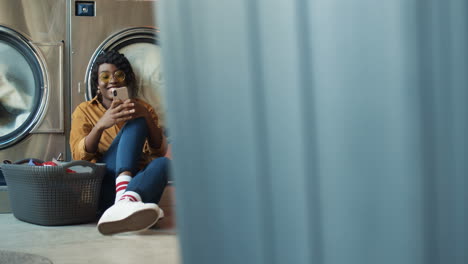 Niña-Sentada-En-El-Piso-Con-Una-Canasta-De-Ropa-Sucia-Y-Tocando-En-El-Teléfono-Inteligente-Mientras-Las-Lavadoras-Trabajan-Hermosa-Mujer-Viendo-Videos-Por-Teléfono-O-Charlando-En-El-Servicio-De-Lavandería