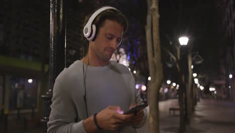 Hombre-Caucásico-Usando-Audífonos,-Escuchando-Música,-Usando-Su-Teléfono-Por-La-Noche