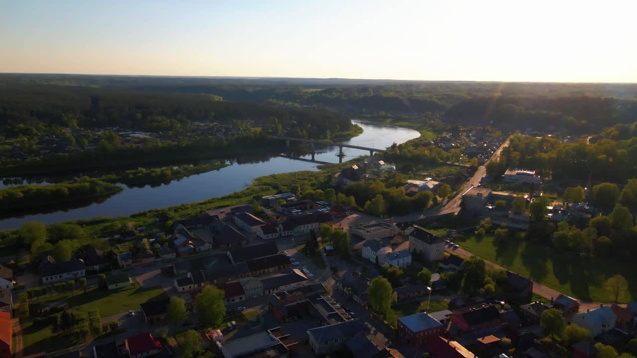 Droneopptak som flyr sidelengs over en liten by ved solnedgang, fanger en elvebro og omkringliggende natur. Opptaket roterer jevnt og avslører hustak og fjern grønn natur i gyllen time-lys.
