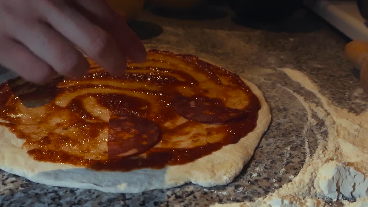 Video al rallentatore ravvicinato di uno/a chef o cuoco/a amatoriale che mette della carne su un impasto per pizza appena steso e fatto a mano con salsa di pomodoro, su un tavolo da cucina con farina bianca intorno