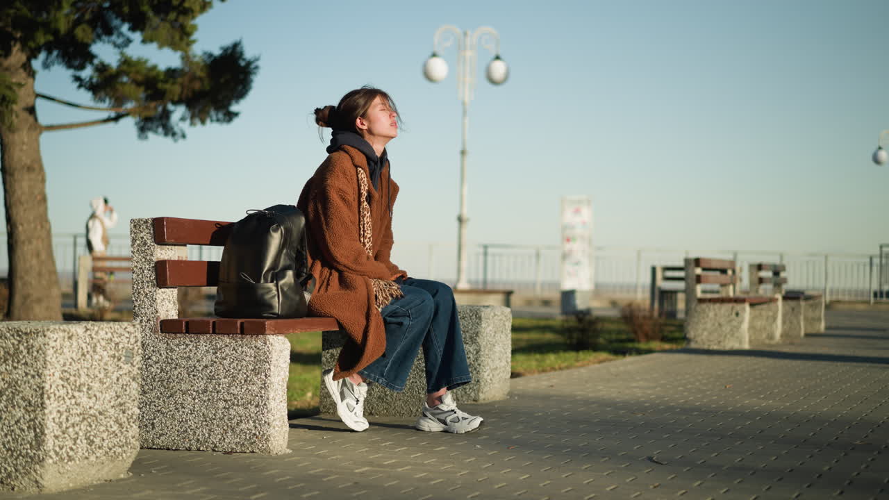 한 소녀가 도시 공원의 벤치에 혼자 앉아, 눈을 감고 생각에 빠져, 갈색 코트, 파란 청바지, 색 신발을 입고, 에 검은색 배을 고 있다.