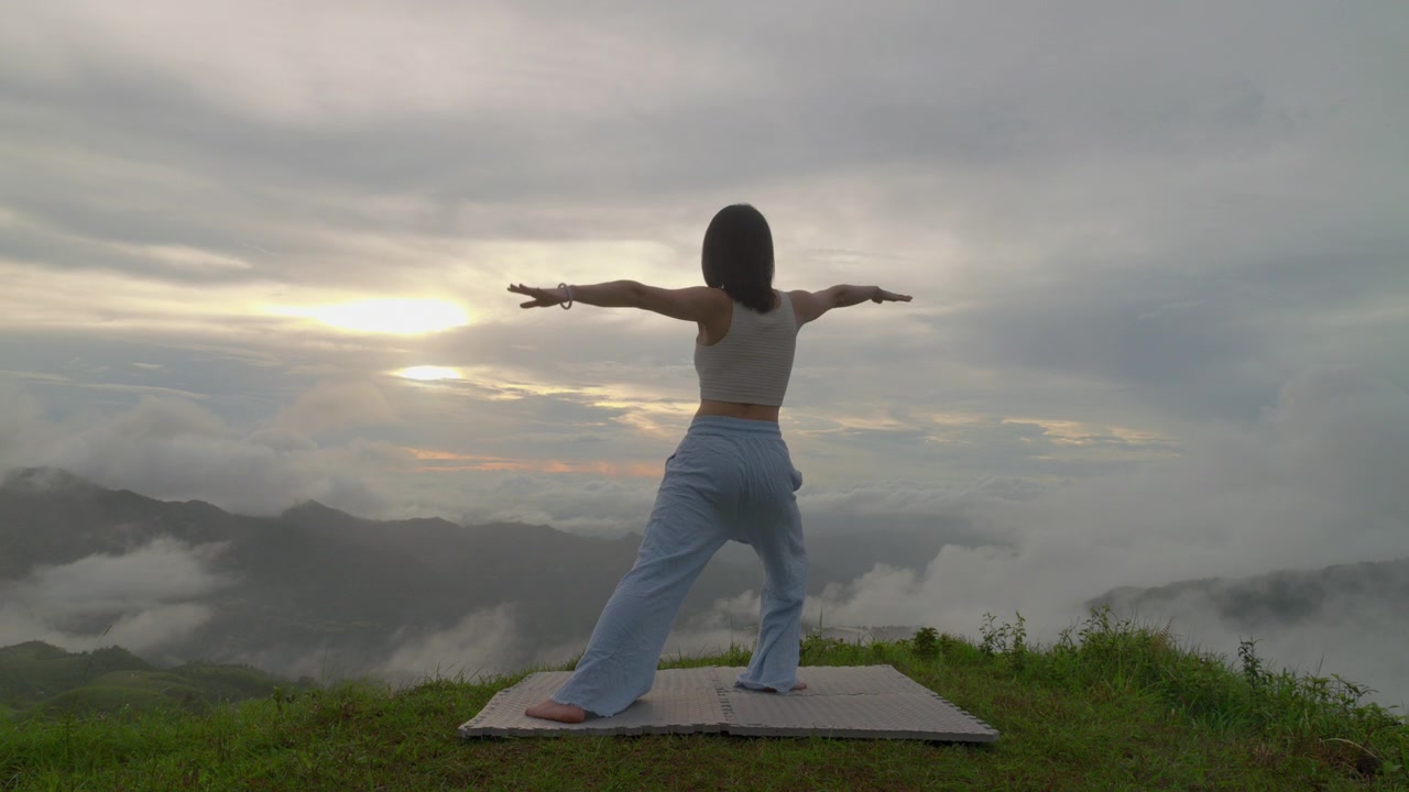 Donna che pratica una posa yoga su un tappetino all'aperto contro uno sfondo di cielo nuvoloso drammatico. Inquadratura ampia