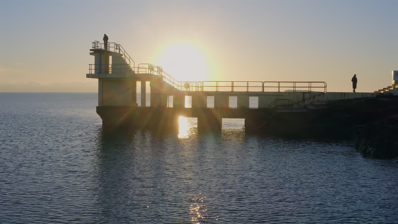 Aerial drone footage gliding over the Galway coast with a backlit diving board at sunset.