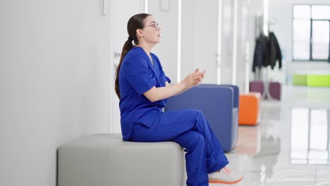 sad girl brunette doctor in round glasses and a blue uniform sits on a soft chair in a modern corridor in the clinic and is sad about failure while working