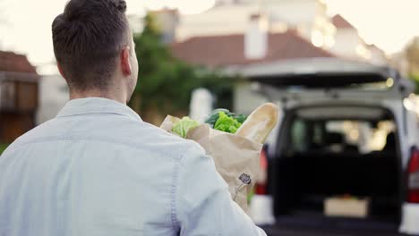 hombre guapo irreconocible yendo al coche llevando una bolsa de papel con comestibles al aire libre