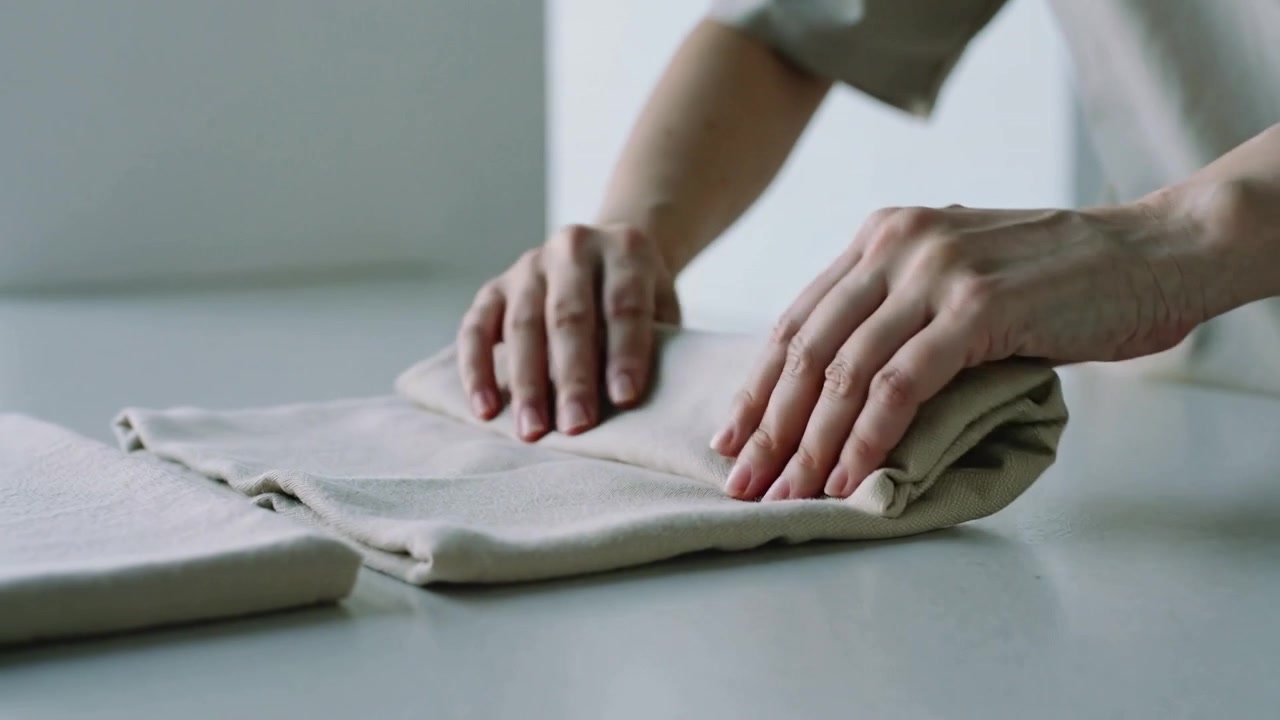 Entering rolled-sleeved hands smoothing and folding cloth on tabletop making neat fold beside linen