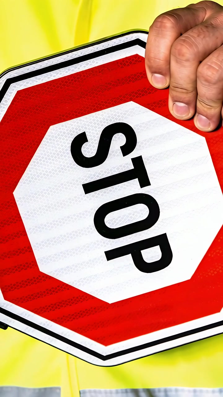 Video verticale: Addetto con giubbotto giallo a bordo strada che ferma il traffico con un segnale di STOP tenuto con la mano destra.