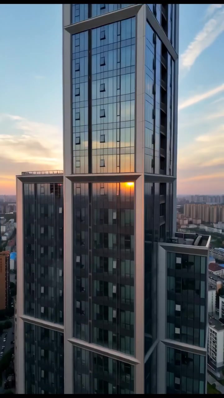 Uma alta torre de escritórios de vidro eleva-se sobre o centro da cidade, capturando reflexos quentes do pôr do sol em sua fachada, com edifícios circundantes, ruas e o céu noturno aberto.