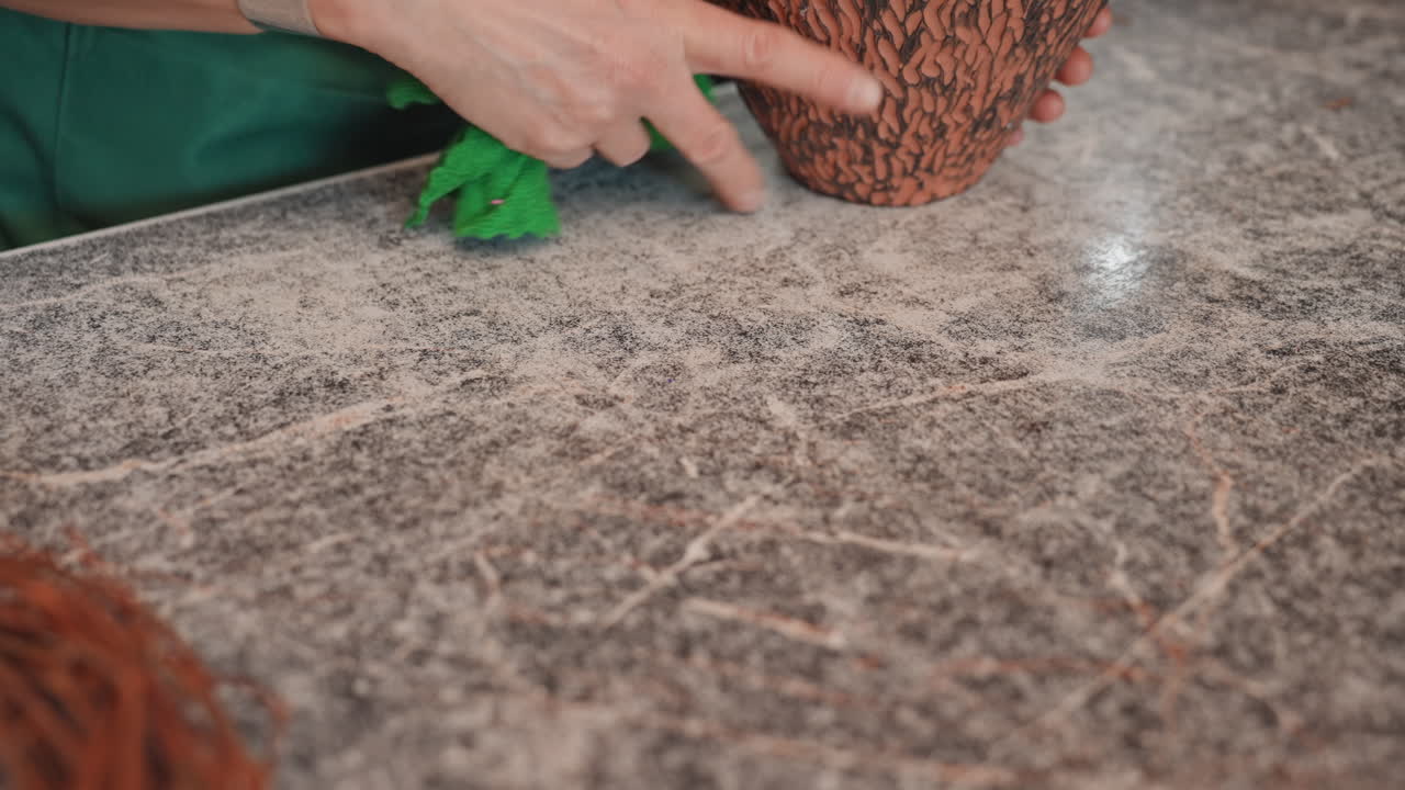 Hands cleaning rustic table with green cloth closeup florist prepping workstation, wiping debris near grapevine wreath and empty pot, textured stone surface, ambient studio light, quiet methodical