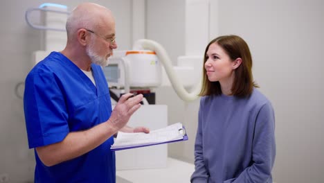 A-confident-elderly-male-doctor-in-glasses-with-a-blue-uniform-communicates-with-a-young-brunette-girl-and-gives-her-advice-on-disease-prevention-during-an-appointment-with-a-doctor-in-a-modern-clinic