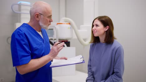 un médico de edad avanzada confiado con gafas con un uniforme azul se comunica con una joven morena y le da consejos sobre la prevención de enfermedades durante una cita con un médico en una clínica moderna