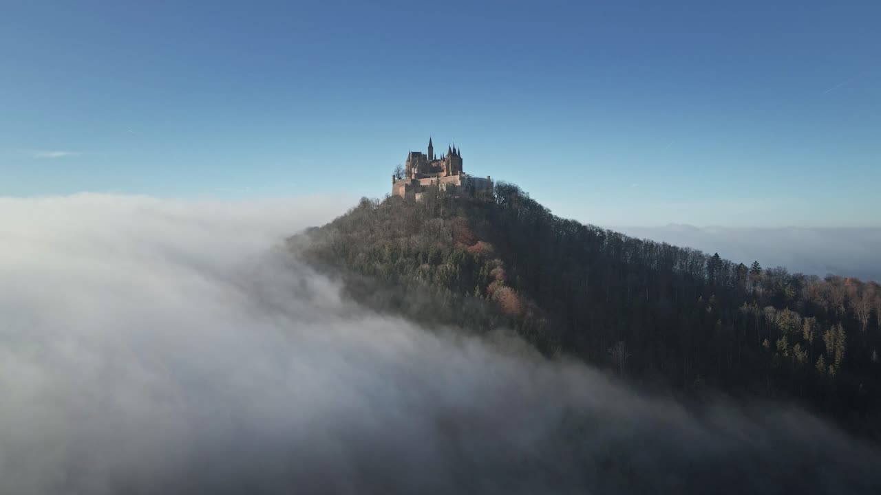 The drone soars further over the clouds, revealing a breathtaking closer view of Hohenzollern Castle as it stands proudly above the mist, creating a dreamy and cinematic landscape.