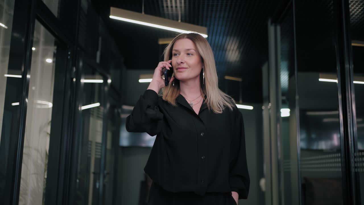A blonde business woman of 30-40 years old in a black shirt walks down the corridor of the office and talks on the phone