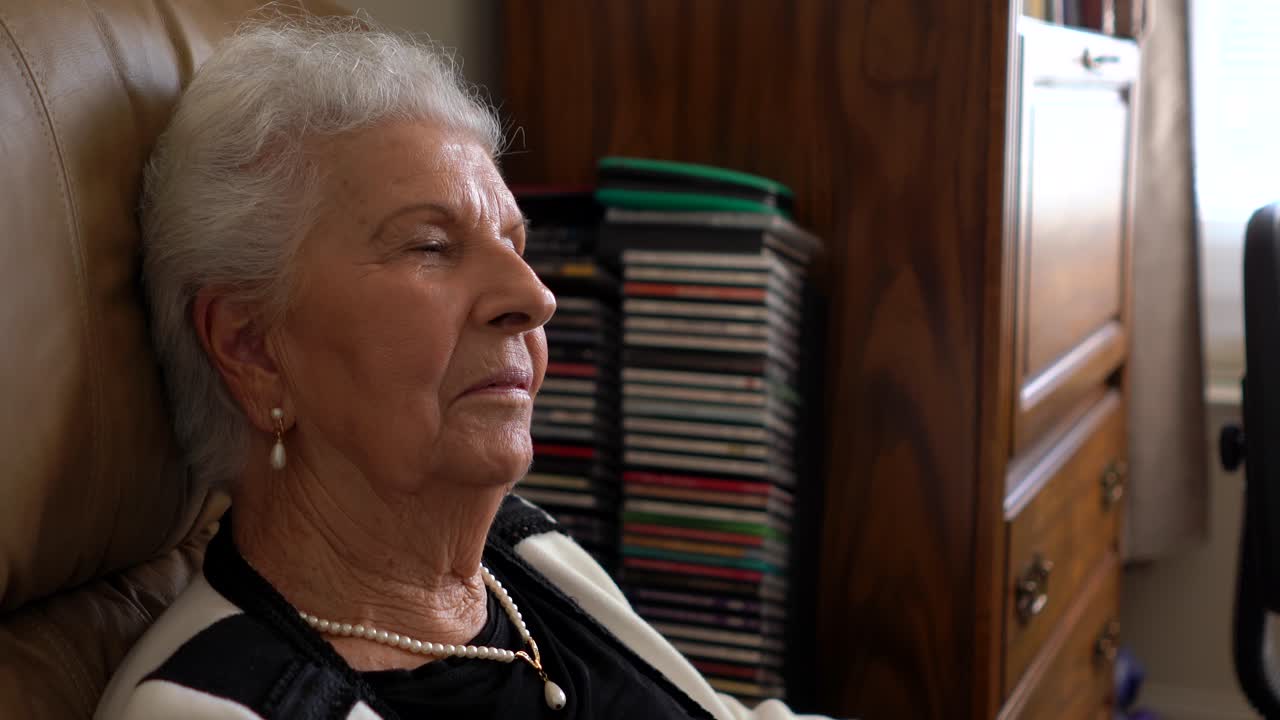 Primo piano di una donna anziana seduta su una sedia comoda, con la testa reclinata all'indietro, che parla dolcemente.