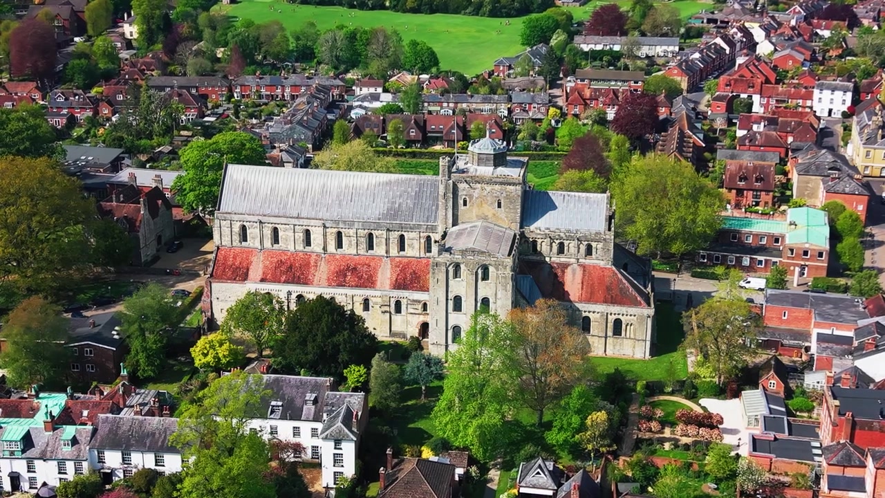 Droneoptagelse fra siden af Romsey Abbey i Hampshire, der viser den historiske klosterkirke, stenarkitektur, røde tagsektioner, omkringliggende hustage, haver, træer og bylandskab.