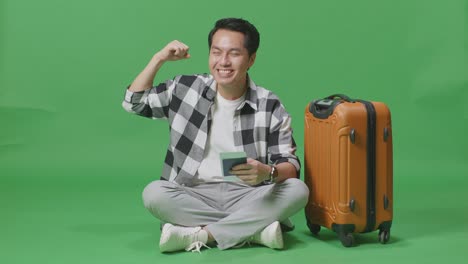 full body of asian male traveler with luggage and passport smiling and flexing his bicep while sitting in the green screen background studio