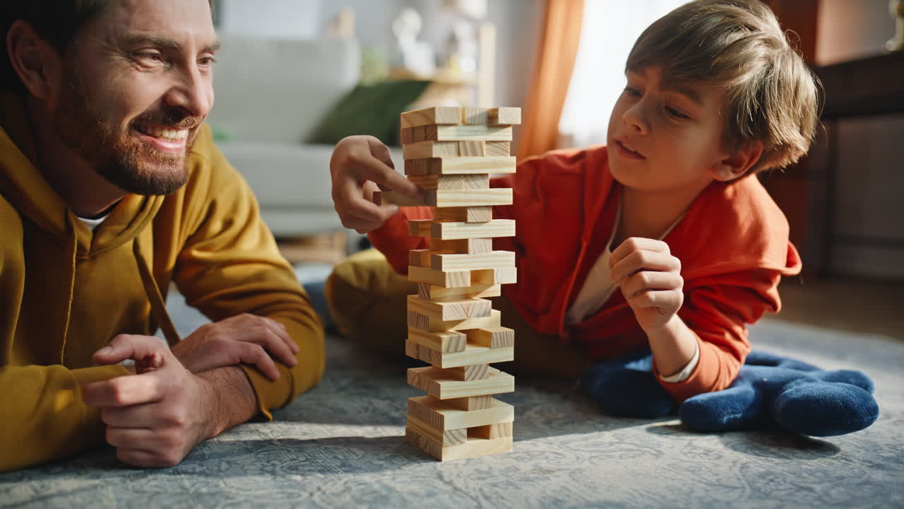 Primo piano: figlio attento che costruisce una torre Jenga con il supporto del papà, sdraiati sul pavimento di casa.