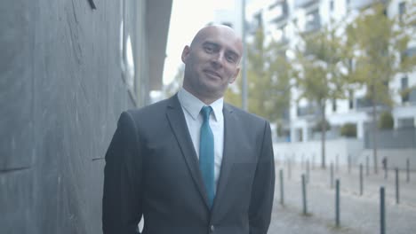 hombre de negocios calvo confiado parado cerca del edificio de oficinas, posando y sonriendo a la cámara