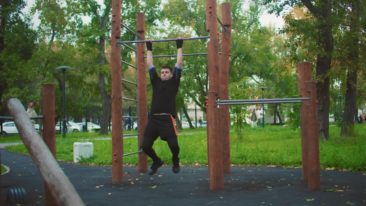 Mann som tar pull-ups på trebarre i parken, svart hettegenser og joggesko, vekslende grep og repetisjoner, fokusert styrketrening og oppvarming, calisthenics-trening med disiplinert form, løvrike trær