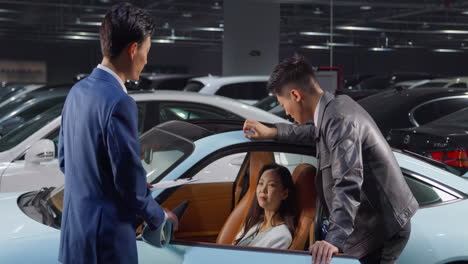 happy couple with the car sales manager in a suit, suggests the client to sit down in the car to look at its interior and quality