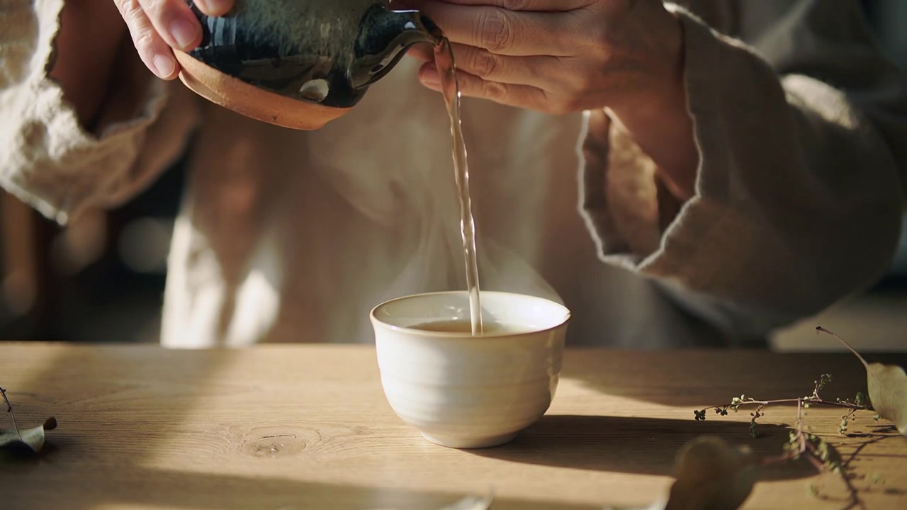 Pouring hands tilting teapot and filling small cup on wooden table, in beige robe, making tea